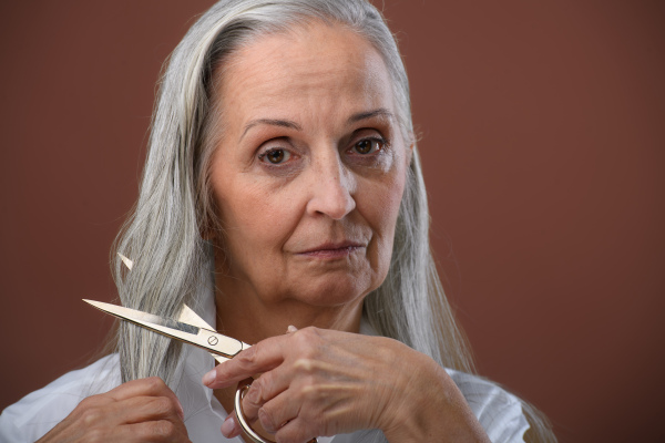 Portrait of unhappy senior woman cutting her hair because of breast cancer. Breast cancer awareness concept.