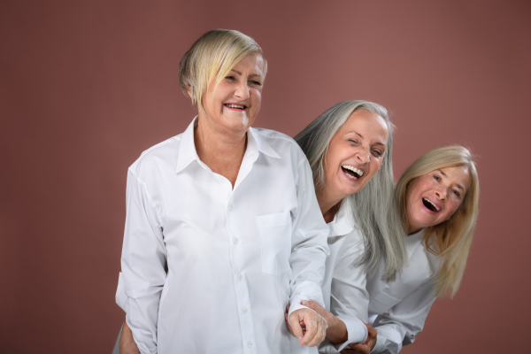 Studio portrait of three beautiful senior women with gray hair in white men's shirt . Elderly funny friends standing behind each other, laughing. Isolated on a brown pink background. Copy space.