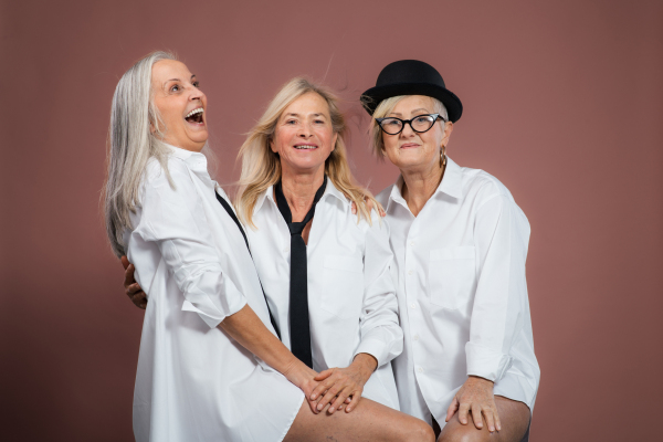 Studio portrait of three beautiful senior women with gray hair in white men's shirt . Elderly funny friends holding each other, looking at camera. Isolated on a brown pink background. Copy space.