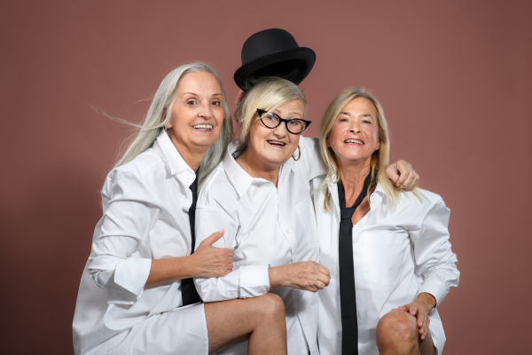 Studio portrait of three beautiful senior women with gray hair in white men's shirt . Elderly funny friends holding each other, looking at camera. Isolated on a brown pink background. Copy space.