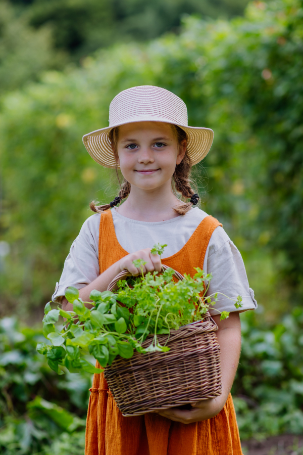 Portrait of a cute little girl in an autumn garden. The young girl in a dress and hat holding a basket full of harvested vegetable and herbs.