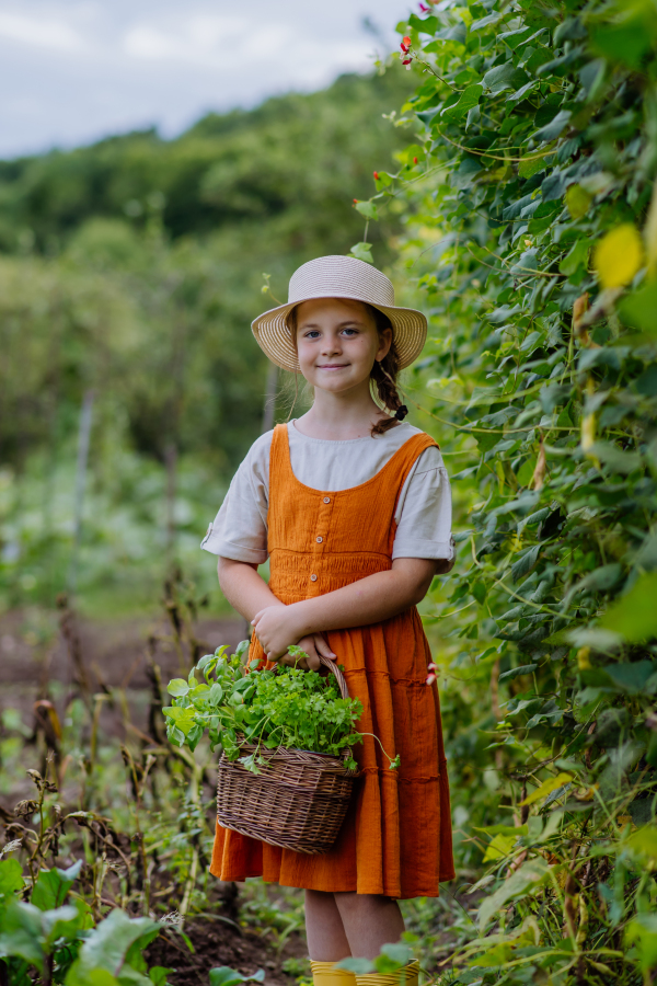 Portrait of a cute little girl in an autumn garden. The young girl in a dress and hat holding a basket full of harvested vegetable and herbs.