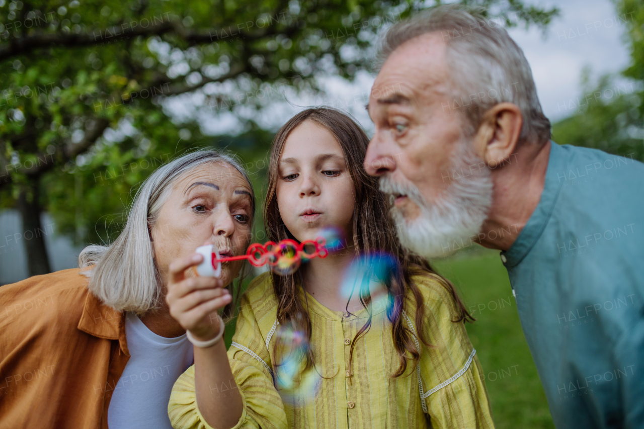 Grandparents and granddaughter are blowing bubbles from a bubble wand. Intergenerational family having fun outdoors in the garden during warm autumn day.