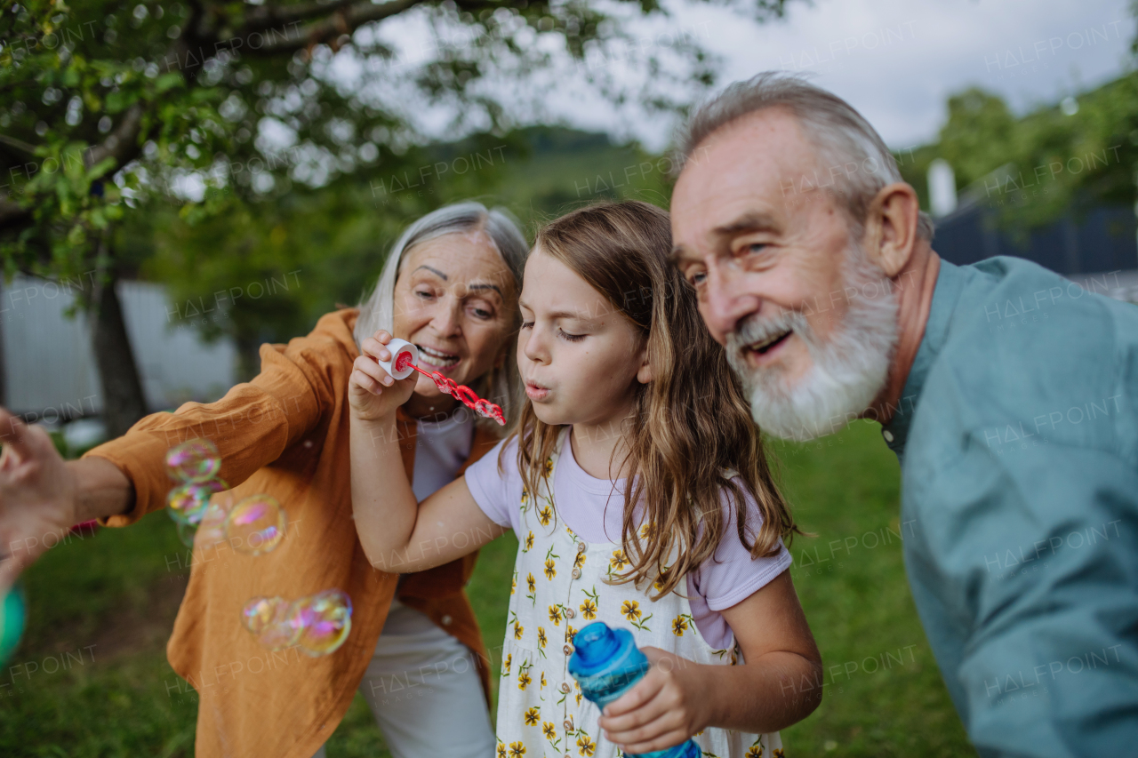 Grandparents and granddaughter are blowing bubbles from a bubble wand. Intergenerational family having fun outdoors in the garden during warm autumn day.