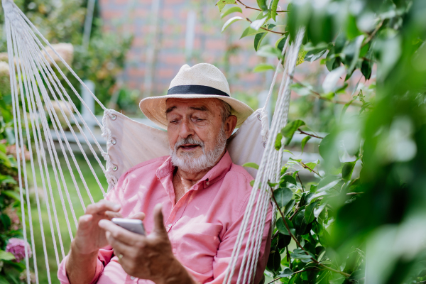 Happy senior man sitting in garden swing and scrolling smartphone.