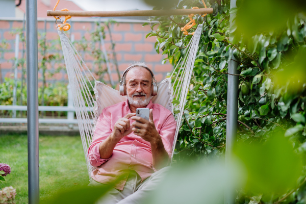 Happy senior man sitting in garden swing with headphones and scrolling smartphone.