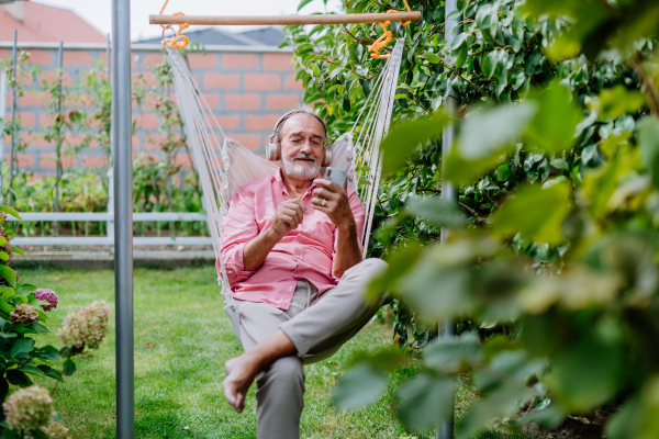 Happy senior man sitting in garden swing, listening music in headphones and scrolling smartphone.