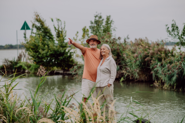 Happy senior couple on walk near the lake during cloudy autumn day, looking at the distance.