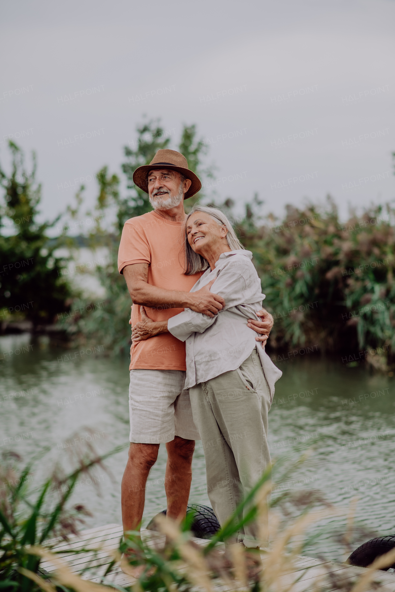 Happy senior couple hugging on walk near the lake during cloudy autumn day.