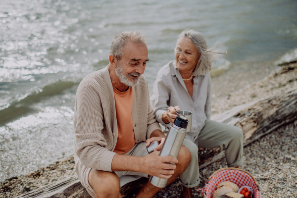 Senior couple sitting, resting and drinking tea from thermos, near the autumn river.