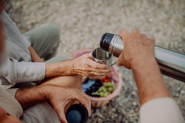 Close-up of senior couple having picnic at beach during autumn day, drinking tea from thermos.