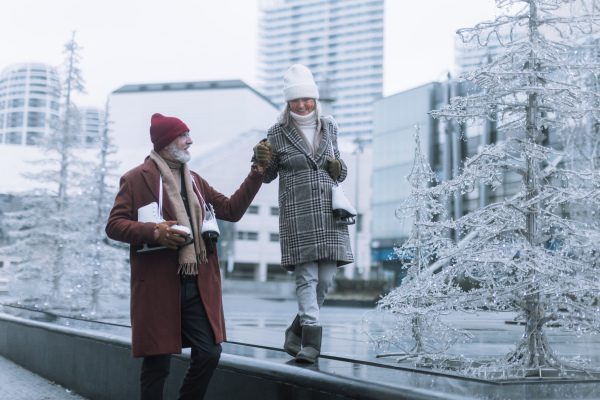 Portrait of seniors in winter at an outdoor ice skating rink. Beautiful elderly woman walking on top of wall holding husband's hand for balance.