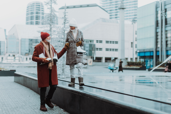 Portrait of seniors in winter at an outdoor ice skating rink. Beautiful elderly woman walking on top of wall holding husband's hand for balance.