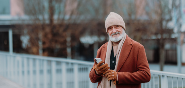 Elegant senior man walking near a river, during cold winter day. Elderly man holding smart phone. Winter banner with copy space.