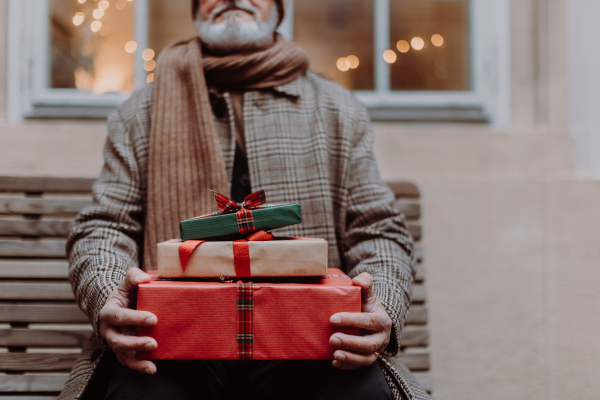 Close-up of senior man sitting on a bench, holding a lot of Christmas gifts in his hands. Christmas shopping spree.