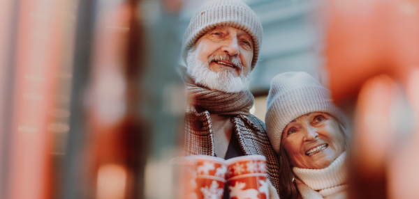 Happy senior couple enjoying outdoor christmas market at the evening, drinking muled wine from christmas mugs. Christmas banner with copy space.