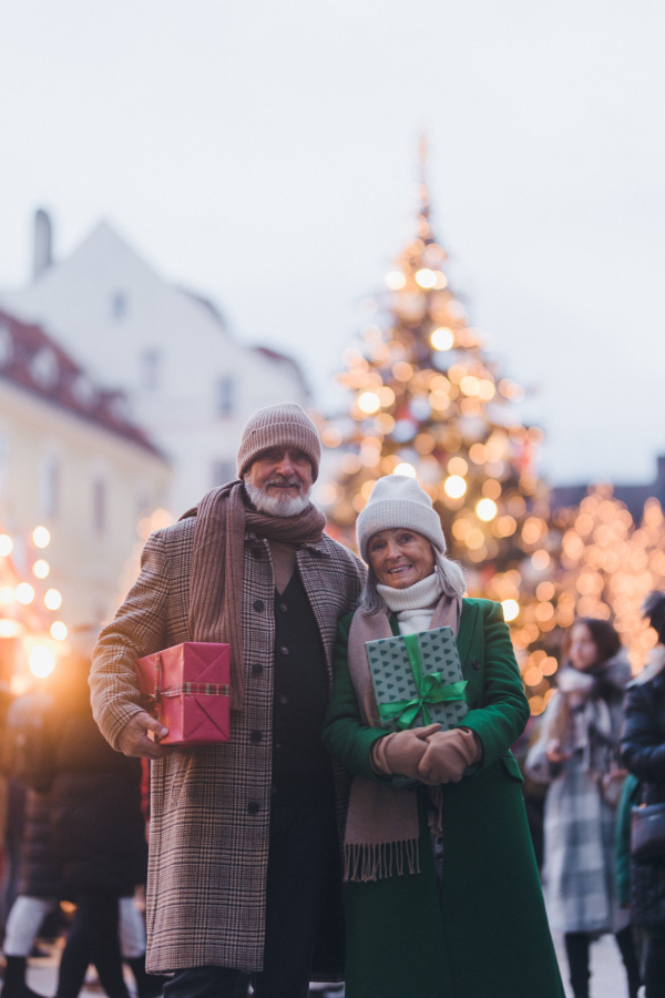 Happy senior couple enjoying outdoor christmas market in the city, buying gifts. Christmas square with a big Christmas tree in the center.