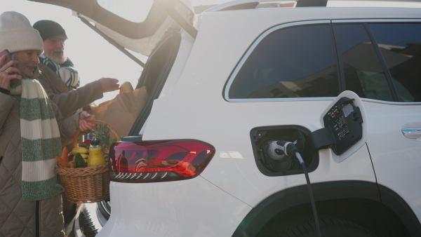 A senior couple charging their electric car, while putting purchase in car trunk.