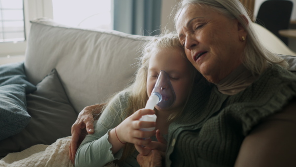 LIttle sick girl using an inhaler during flu while her grandmother comforting her.