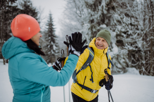 Senior couple skiing together in the middle of snowy forest, having break and giving high five.