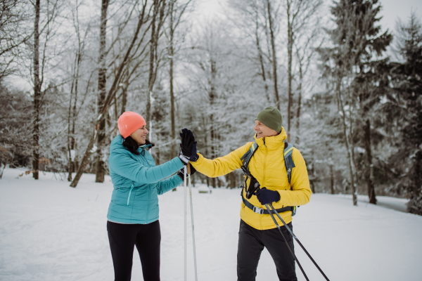 Senior couple skiing together in the middle of snowy forest, having break and giving high five.