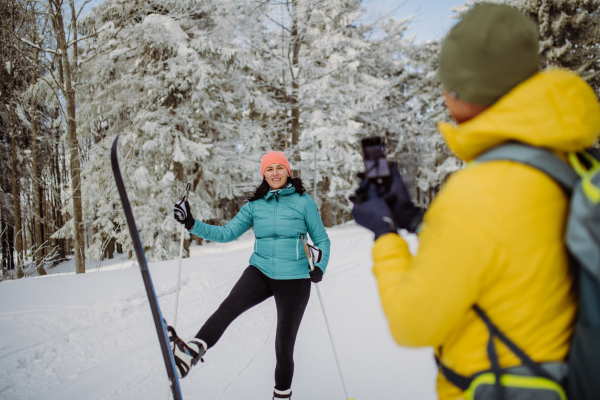 Senior man taking photo of his wife posing with skis in winter forest.