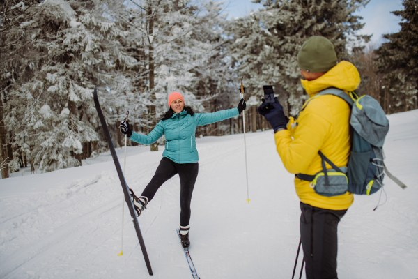 Senior man taking photo of his wife posing with skis in winter forest.