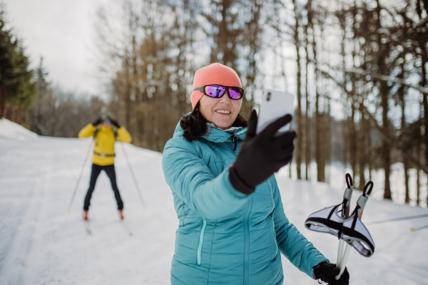 Senior woman taking selfie with her husband in background during cross country skiing in snowy nature.