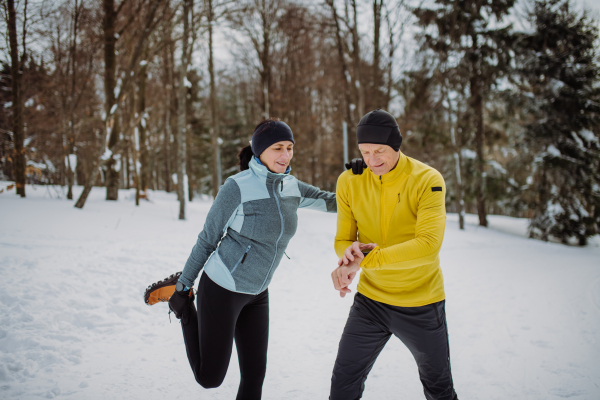 Senior couple warming-up and checking smartwatch in forest before winter hike.