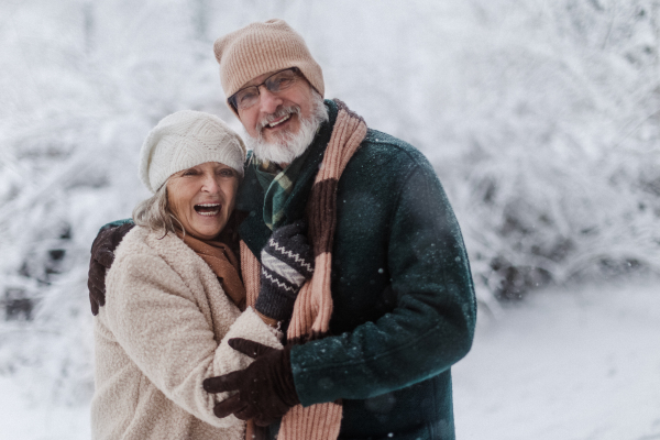 Waist up portrait of elegant senior couple walking in the snowy park, during cold winter snowy day. Elderly couple spending winter vacation in the mountains. Wintry landscape.