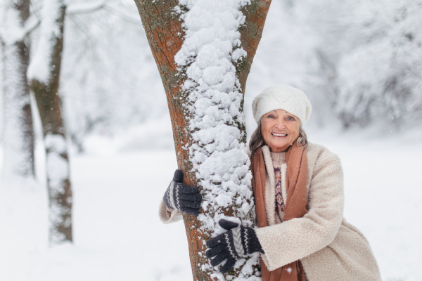 Waist up portrait of elegant senior woman standing by a tree, during cold winter snowy day. Elderly woman spending winter vacation in the mountains. Wintry christmas landscape.