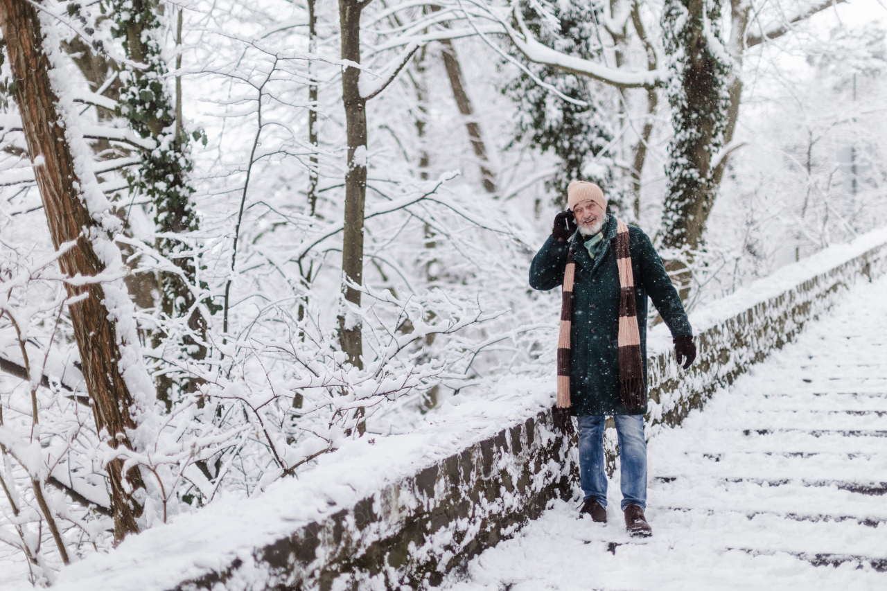 Elegant senior man calling to someone during walk in the snowy park. Elderly man spending winter vacation in the mountains. Wintry landscape.