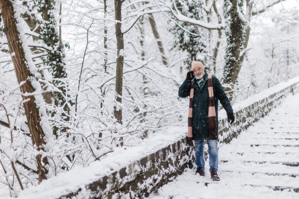 Elegant senior man calling to someone during walk in the snowy park. Elderly man spending winter vacation in the mountains. Wintry landscape.