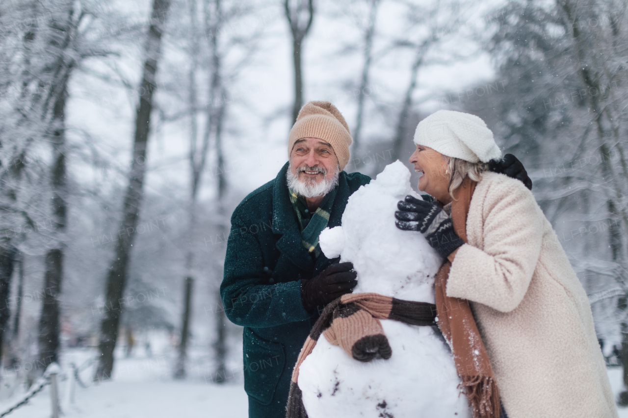 Elegant senior couple building snowman during cold winter snowy day. Elderly couple spending winter vacation in the mountains. Wintry christmas landscape.