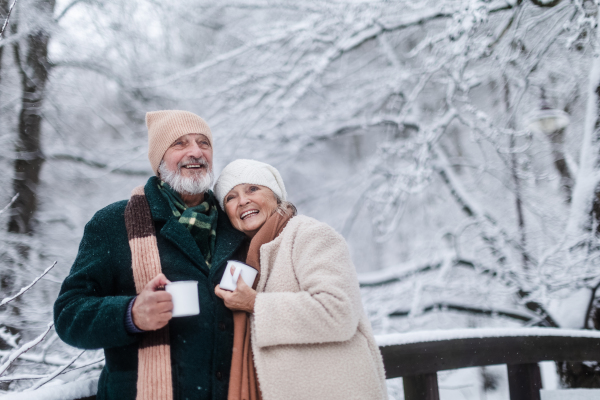 Elegant senior couple having hot tea outdoors, during cold winter snowy day. Elderly couple spending winter vacation in the mountains. Wintry landscape.