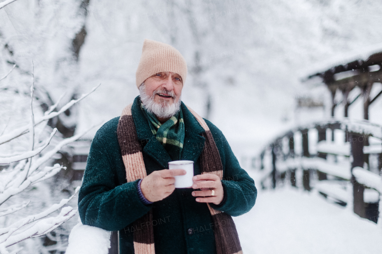 Portrait of elegant senior man drinking hot tea outdoors, during cold winter snowy day. Elderly man spending winter vacation in the mountains. Wintry landscape.