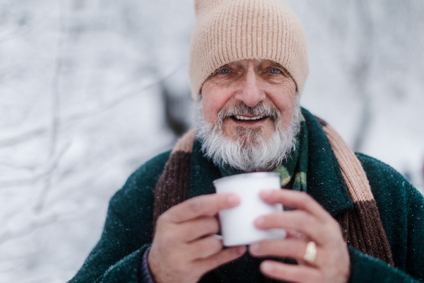 Portrait of elegant senior man drinking hot tea outdoors, during cold winter snowy day. Elderly man spending winter vacation in the mountains. Wintry landscape.