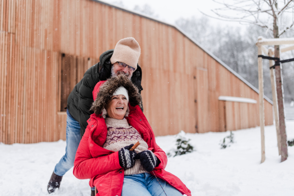 Senior couple having fun during cold winter day, sledding down the hill. Senior woman in red coat on sled. Elderly couple spending winter vacation in the mountains. Wintry landscape.