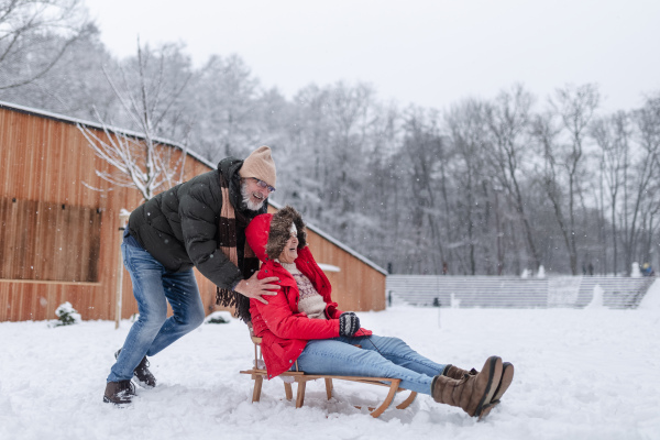 Senior couple having fun during cold winter day, sledding down the hill. Senior woman in red coat on sled. Elderly couple spending winter vacation in the mountains. Wintry landscape.