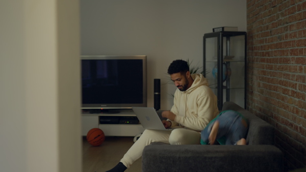 Multiracial father and his son having fun in their apartment. Father working on laptop.