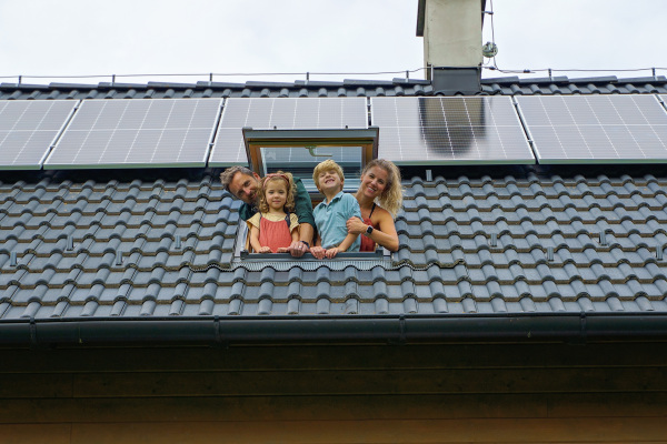 Happy family leaning out from skylight window in their house with solar panels on the roof. Alternative energy, saving resources and sustainable lifestyle concept.