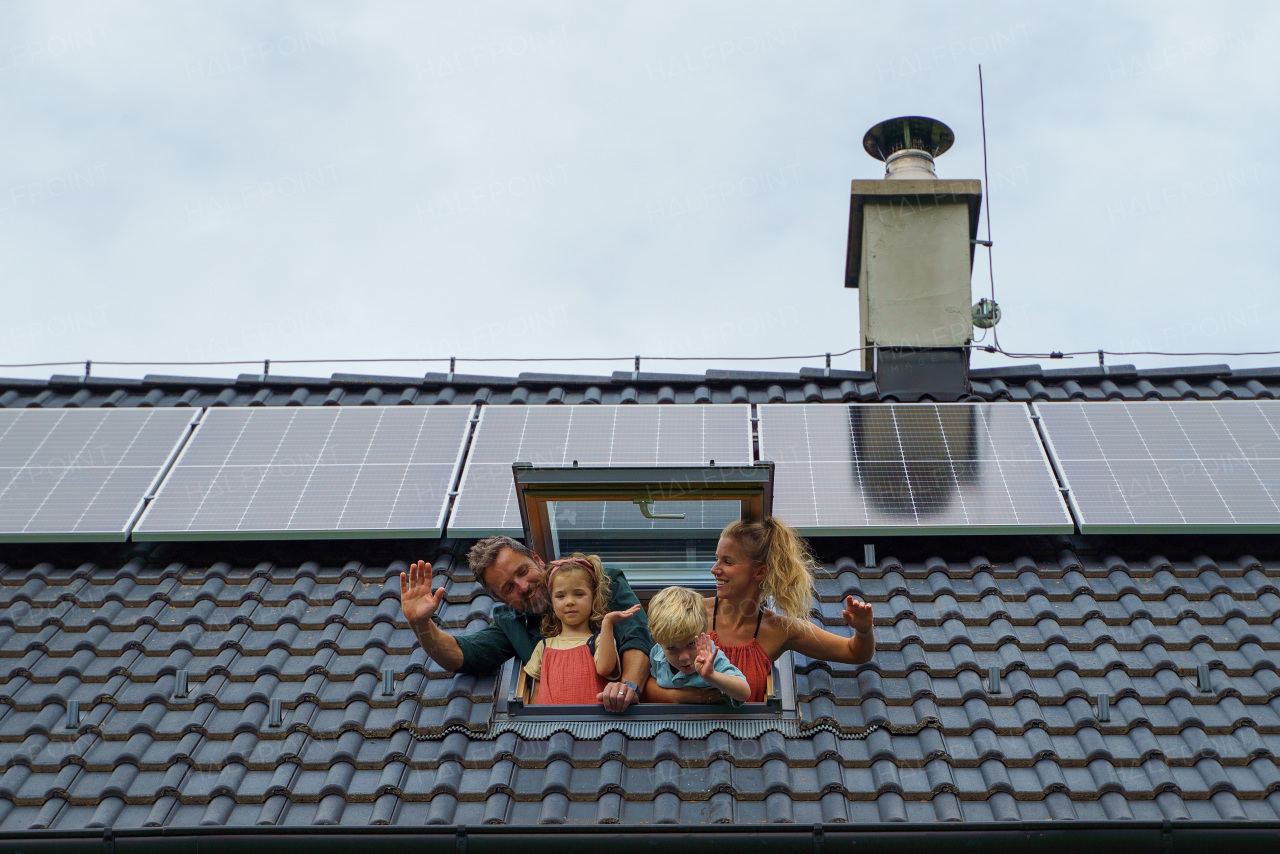 Happy family waving from skylight window in their house with solar panels on the roof. Alternative energy, saving resources and sustainable lifestyle concept.