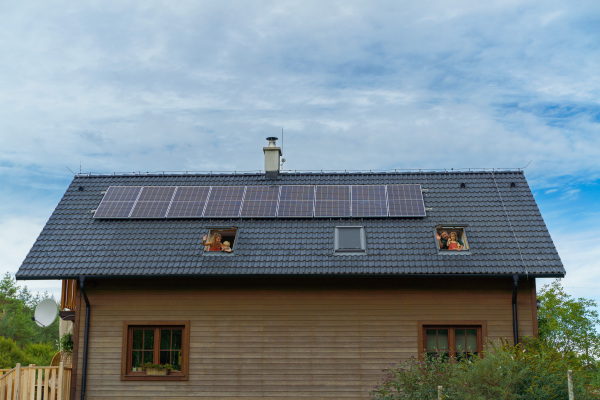 Happy family waving from skylight windows in their house with solar panels on the roof. Alternative energy, saving resources and sustainable lifestyle concept.