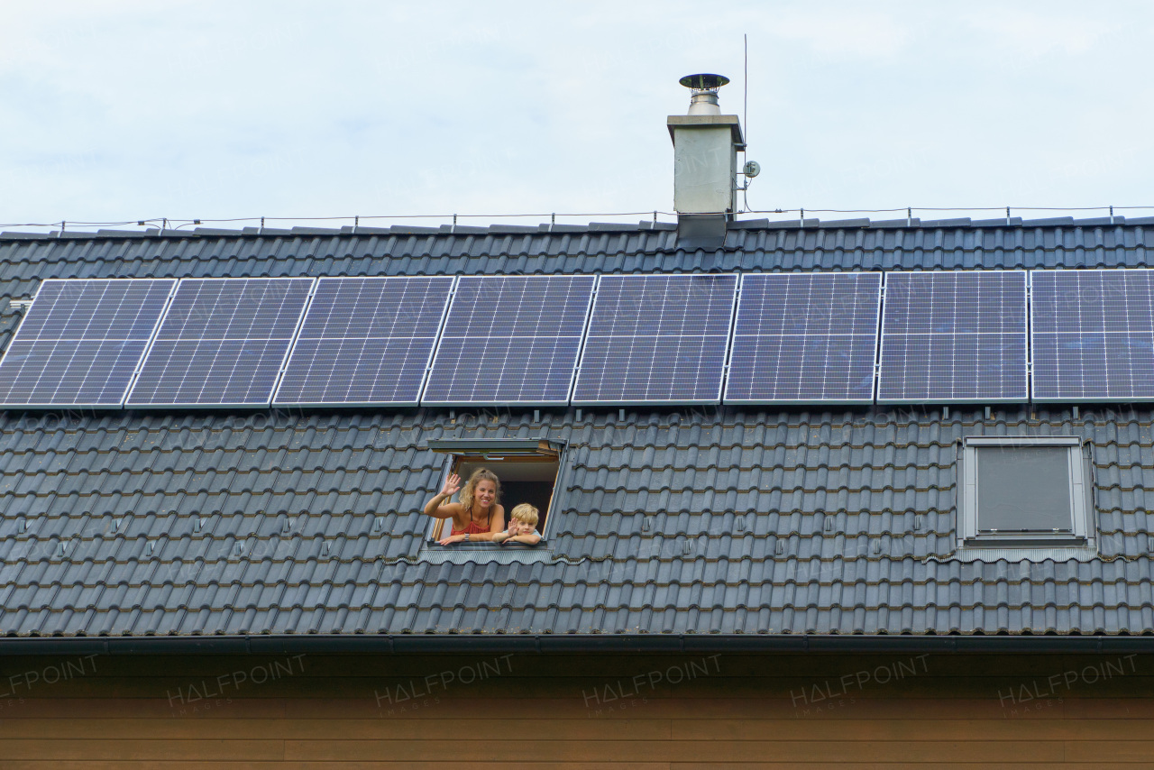 Happy mother and son waving from skylight windows in their house with solar panels on the roof. Alternative energy, saving resources and sustainable lifestyle concept.