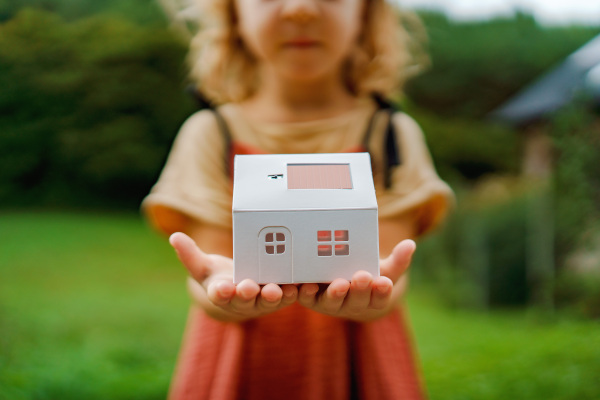 Close up of little girl holding paper model of house with the solar panels.Alternative energy, saving resources and sustainable lifestyle concept.