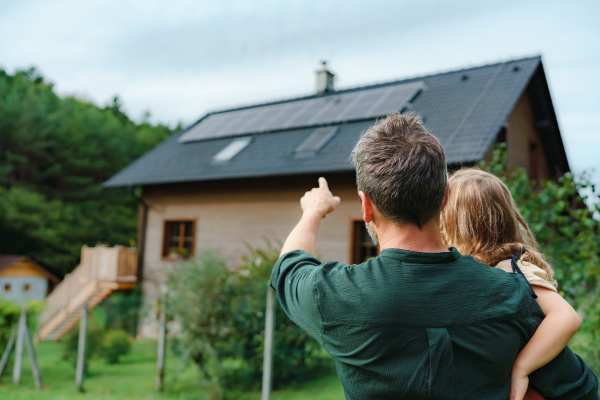 Rear view of dad holding her little girl in arms and showing at their house with solar panels.Alternative energy, saving resources and sustainable lifestyle concept.