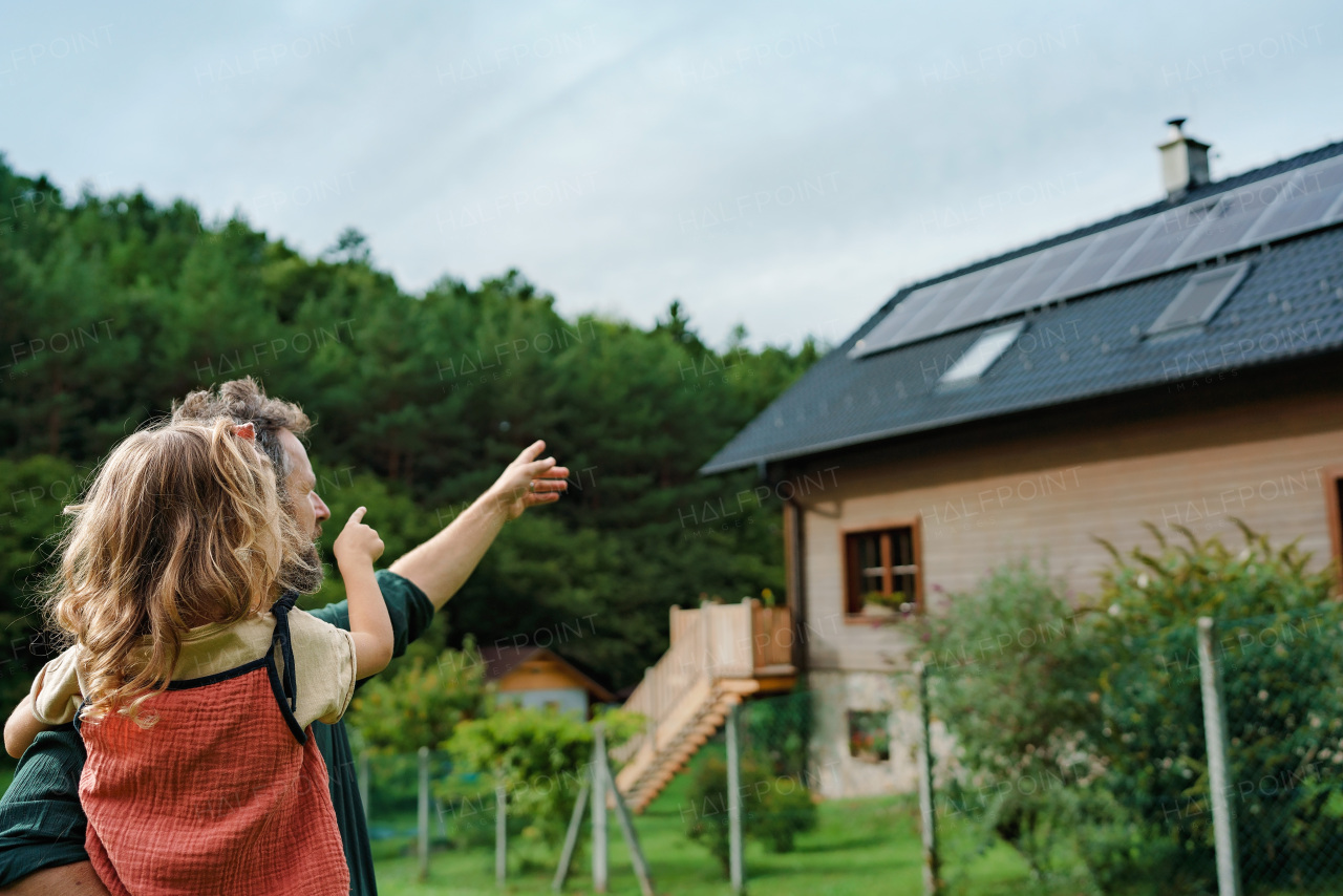 Rear view of dad holding her little girl in arms and showing at their house with solar panels.Alternative energy, saving resources and sustainable lifestyle concept.