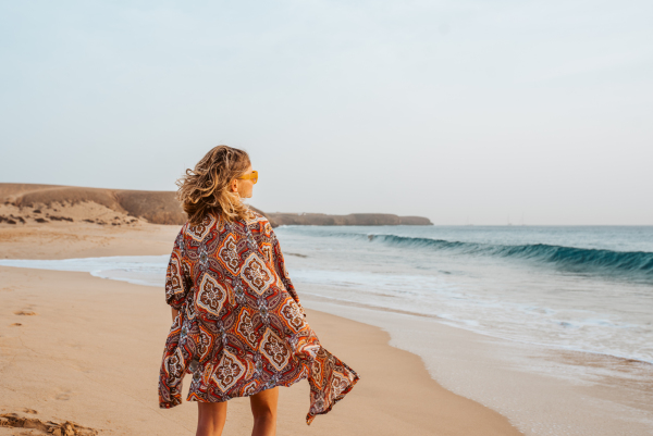 Rear view of a beautiful slim woman standing on sand beach. Full body shot of barefoot woman looking at sea. Concept of beach summer vacation.