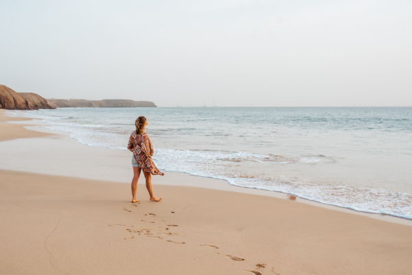 Rear view of a beautiful slim woman standing on sand beach. Full body shot of barefoot woman looking at sea. Concept of beach summer vacation.