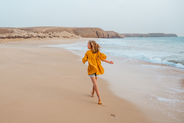 Young girl on the beach. Rear view of blonde girl enjoying sandy beach, looking at crystalline sea in Canary Islands. Concept of beach summer vacation with kids.